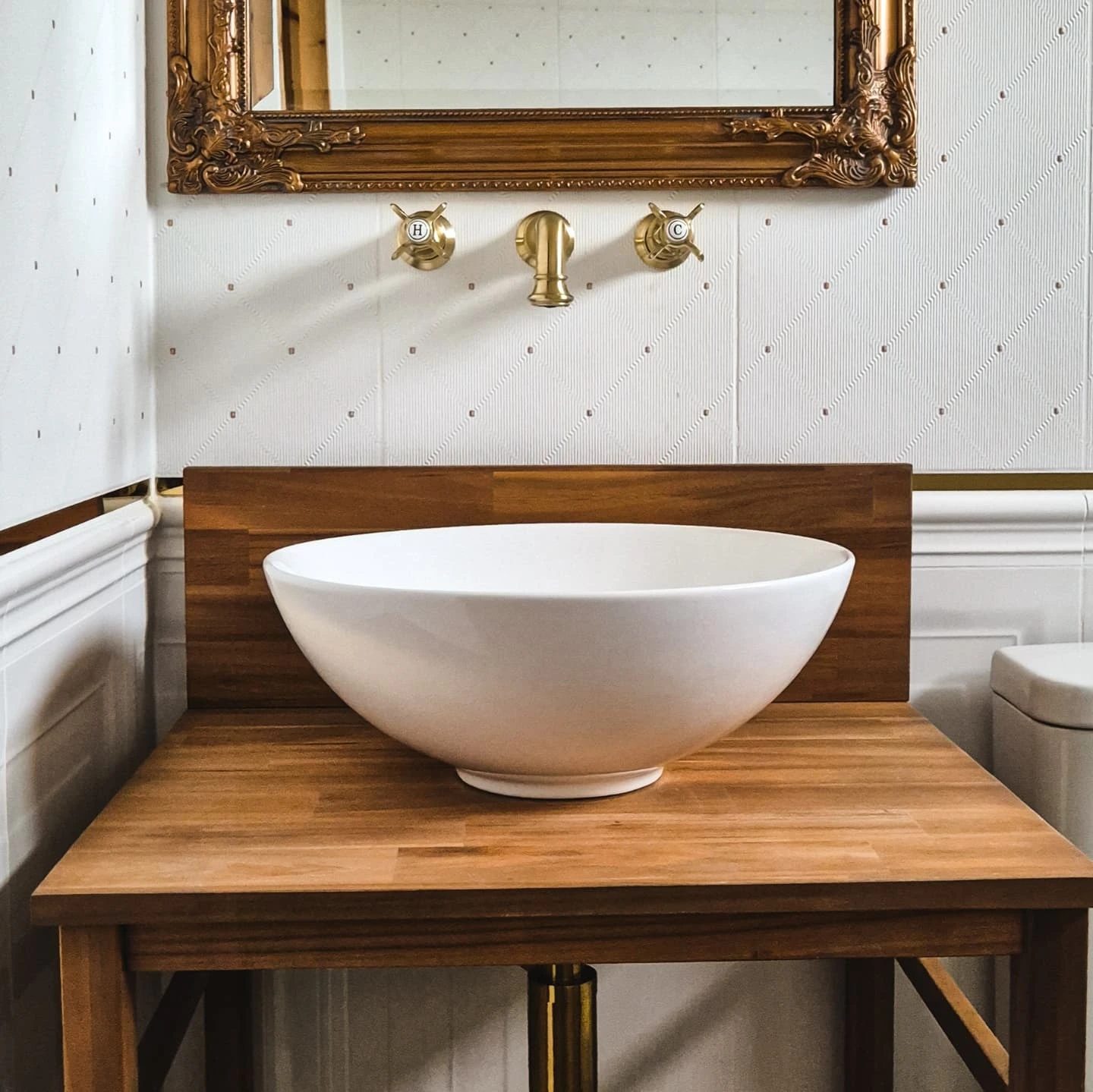 A timber vanity stand with a white basin and brass taps under a gilt mirror